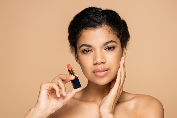 young african american woman with bare shoulders holding lipstick tube isolated on beige  - Fotografie, Obrázek