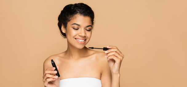happy african american woman with bare shoulders holding mascara tube and brush isolated on beige, banner - Zdjęcie, obraz