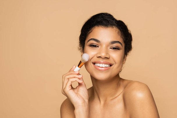 boldog afro-amerikai nő csupasz vállak alkalmazása arc por kozmetikai kefe izolált bézs  - Fotó, kép
