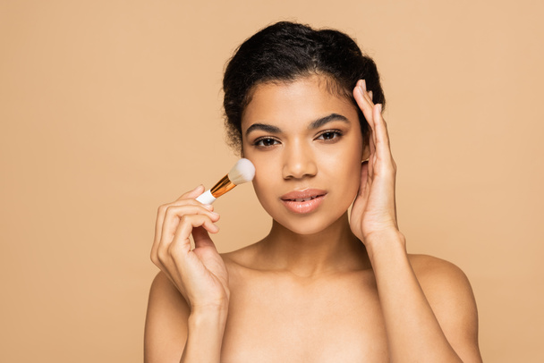 young african american woman with bare shoulders applying face powder with cosmetic brush isolated on beige  - 写真・画像