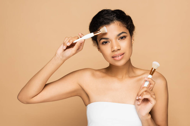 young african american woman with bare shoulders holding cosmetic brushes isolated on beige  - Foto, Imagem