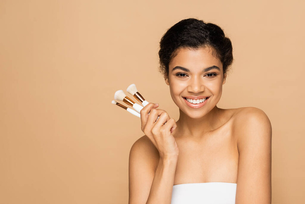 boldog fiatal afro-amerikai nő csupasz vállak gazdaság kozmetikai kefék elszigetelt bézs  - Fotó, kép