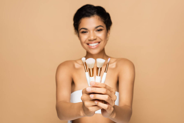 blurred and cheerful african american woman with bare shoulders holding cosmetic brushes isolated on beige  - Photo, Image