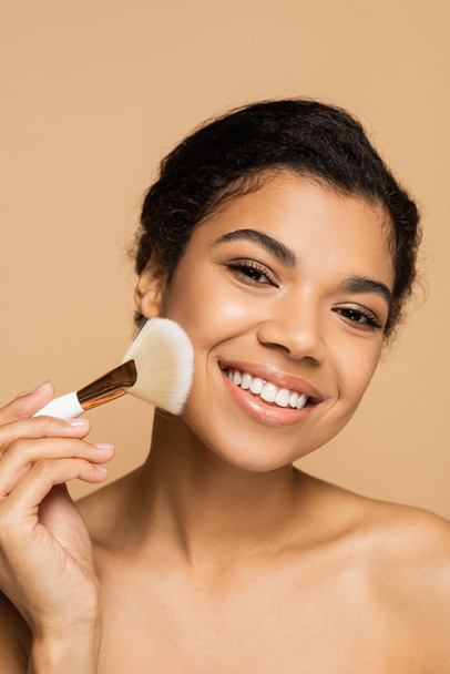 mosolygós afro-amerikai nő alkalmazása arc por kozmetikai kefe izolált bézs  - Fotó, kép