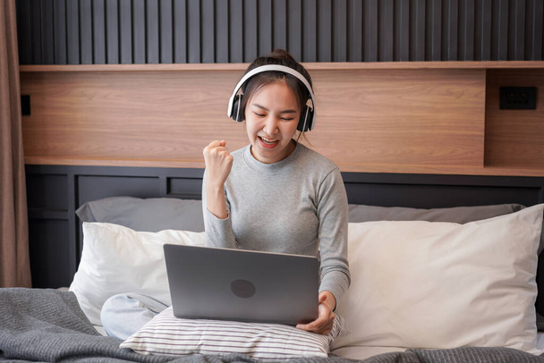 Young asian woman with headphone showing yes gesture with hand after receive email good news while sitting to working and listening to music from laptop on the bed in home bedroom - Foto, afbeelding