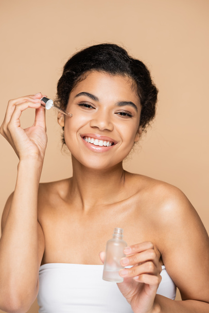 happy african american woman with bare shoulders applying serum with pipette isolated on beige  - Fotografie, Obrázek