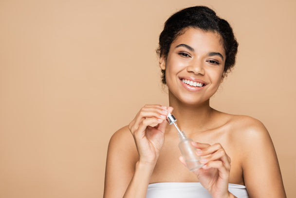 cheerful african american woman with bare shoulders holding serum bottle and pipette isolated on beige  - Foto, Imagem