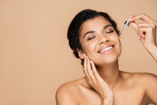 happy african american woman with bare shoulders applying serum isolated on beige  - Foto, Imagem