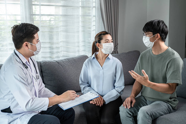 Man doctor in medical face mask to conversation with young couple while he holding document to discussion about result heath examination and giving advice treatment to both during visit at home - Foto, Bild