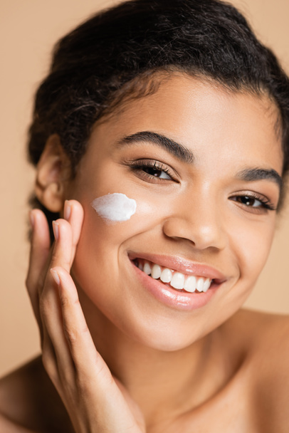 smiling african american woman applying face cream on cheek isolated on beige  - Photo, image
