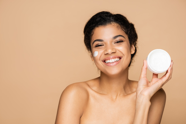 smiling african american woman with face cream on cheeks holding container isolated on beige - Фото, изображение