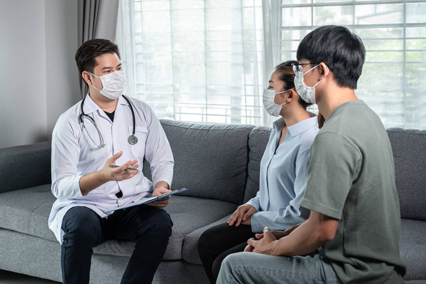 Man doctor in medical face mask holding clipboard to discussion about symptoms and disease after using stethoscope with patient and giving advice treatment for young couple during medical help at home - Fotografie, Obrázek