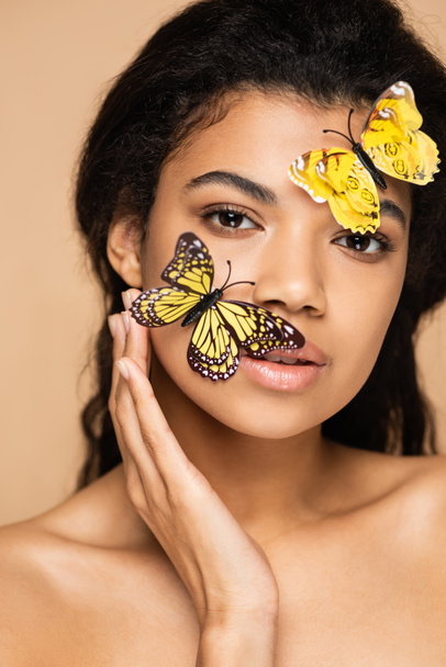 fiatal afro-amerikai nő sárga dekoratív pillangók az arcon nézi kamera elszigetelt bézs - Fotó, kép