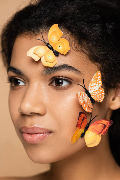 κοντινό πλάνο της νεαρής Αφροαμερικανής γυναίκας με πεταλούδες στο πρόσωπο κοιτάζοντας μακριά απομονωμένη στο μπεζ - Φωτογραφία, εικόνα