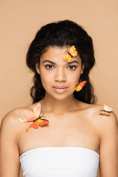 pretty african american woman with orange butterflies on face and bare shoulders looking at camera isolated on beige - Фото, изображение
