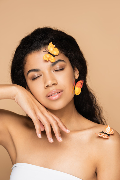mooie Afro-Amerikaanse vrouw met gesloten ogen, oranje vlinders op gezicht en blote schouders geïsoleerd op beige - Foto, afbeelding