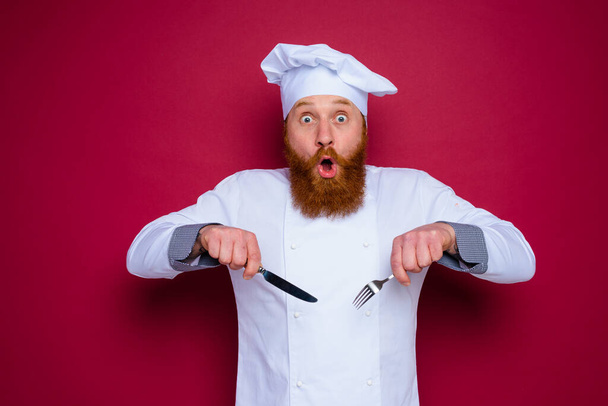 verblüffter Koch mit Bart und roter Schürze hält Besteck in der Hand - Foto, Bild