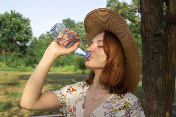 Una joven bebe agua de una botella en la naturaleza. Agua potable pura en una botella de plástico. Apagando la sed mientras camina - Foto, Imagen