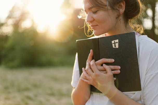 Christian woman holds bible in her hands. Reading the Holy Bible in a field during beautiful sunset. - Photo, Image