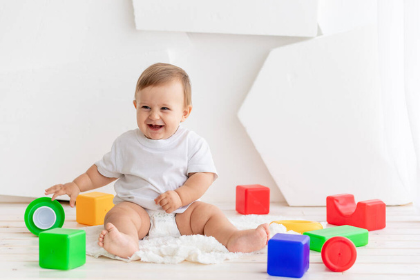 gelukkig baby zes maanden oud in een wit t-shirt en luiers spelen thuis op een mat in een heldere kamer met felgekleurde kubussen - Foto, afbeelding