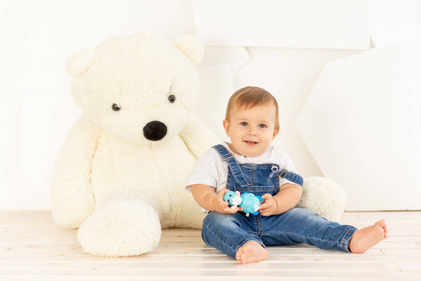 uma criança pequena de seis meses de idade em jeans azuis brinca em casa perto de um grande urso de pelúcia - Foto, Imagem