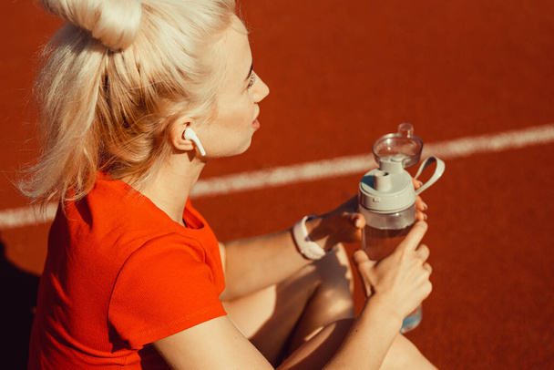 młoda piękna blondynka siedzi na torze joggingowym z butelką wody w rękach - Zdjęcie, obraz