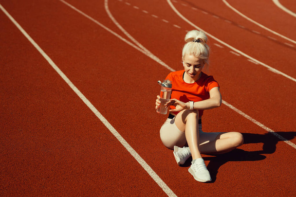 mladá krásná blondýna sedí na běžecké dráze s lahví vody a dívá se na její hodinky - Fotografie, Obrázek
