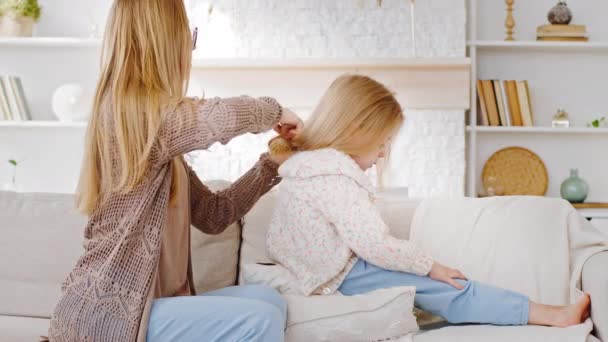 Young family caring caucasian mother woman babysitter mommy sits on sofa with daughter little child toddler preschool girl combing hair of kid doing haircut using comb daily morning beauty procedures - Footage, Video