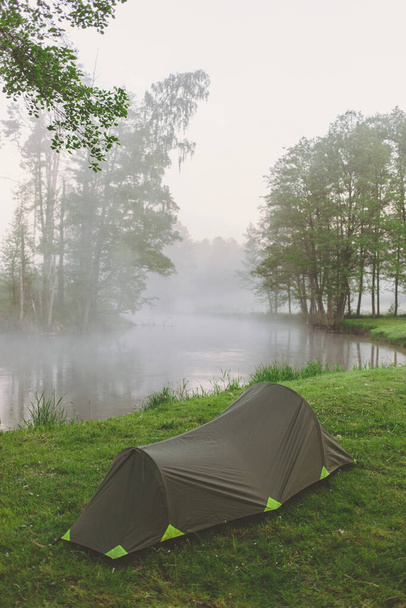 Sisli bir sabahta, nehrin yanındaki ormanda tek yeşil çadır. - Fotoğraf, Görsel