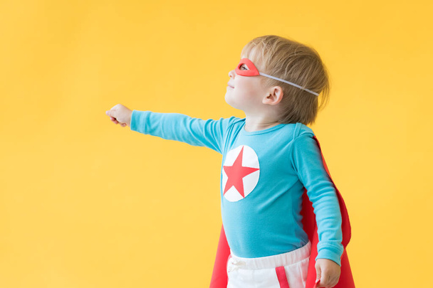 Superhero child against yellow paper background. Super hero kid wearing red mask and cape. Childhood dream and imagination concept - 写真・画像