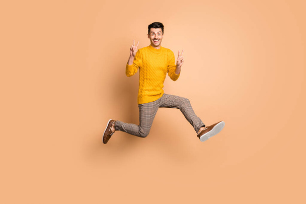 Full length photo portrait of funny man making two v-signs jumping up isolated on pastel beige colored background - Фото, изображение