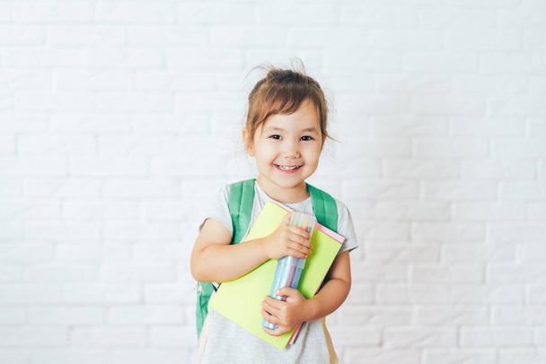 Dziecko z zielonym plecakiem trzymające biurową papeterię przed białym tłem. - Zdjęcie, obraz