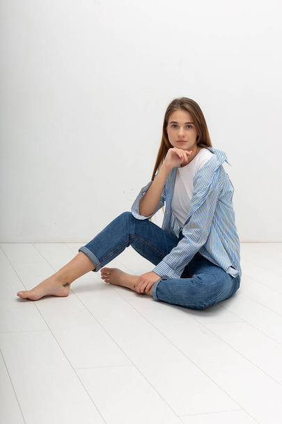 молодая белая симпатичная девушка с длинными волосами в рубашке, синие джинсы сидят в студии - Фото, изображение