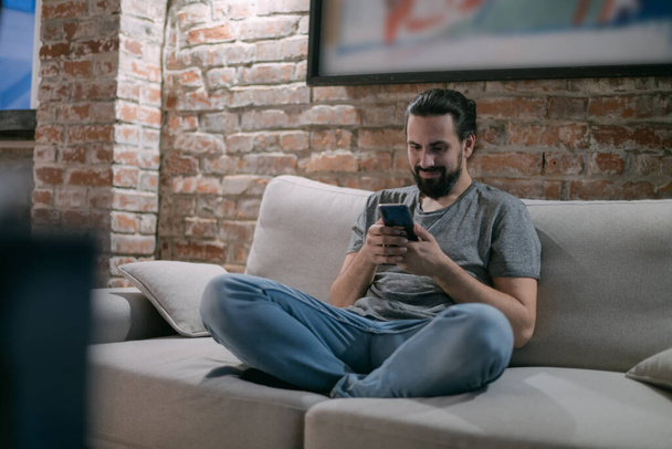 A young man is sitting on the couch with the phone in the evening. A handsome guy is resting, chatting online in a modern, stylish house in the late evening. - Fotoğraf, Görsel