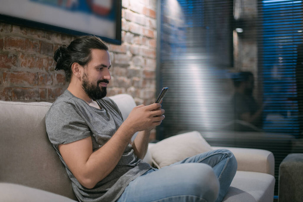 Ein junger Mann sitzt abends mit dem Handy auf der Couch. Ein gutaussehender Kerl ruht sich aus und plaudert am späten Abend online in einem modernen, stilvollen Haus. - Foto, Bild