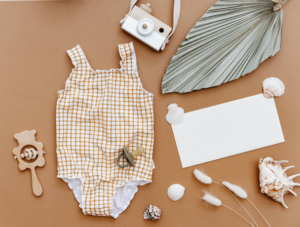 Cute retro swimsuit for baby girl with seashells and beach accessories on brown background. Summer holiday concept. Top view, flat lay  - Photo, Image