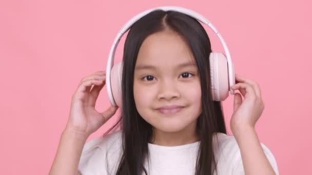 Χαριτωμένο κοριτσάκι από την Ασία με ασύρματα ακουστικά ακούγοντας δυναμική μουσική, χορεύοντας και χαμογελώντας στην κάμερα - Πλάνα, βίντεο