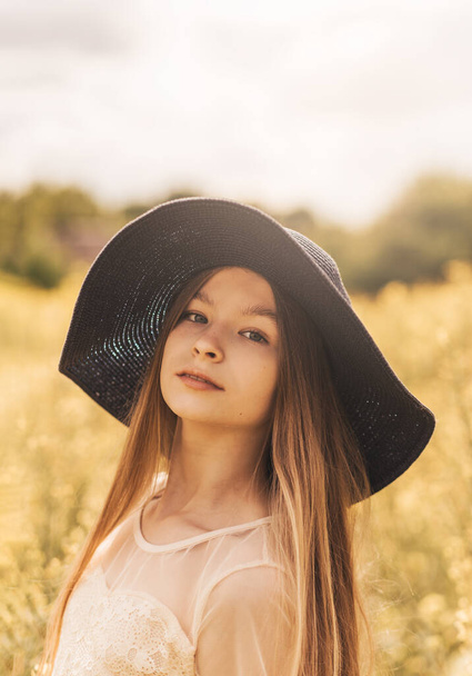 Portrait of a beautiful young girl in a hat against the background of a rapeseed field. Vertical photo - 写真・画像