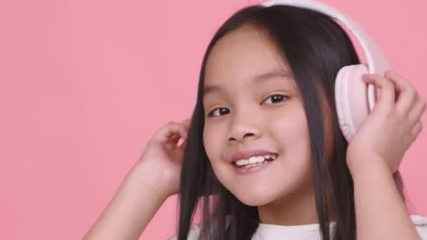 Amante de la música. Alegre niña asiática escuchando música en grandes auriculares inalámbricos rosa, sonriendo a la cámara - Metraje, vídeo