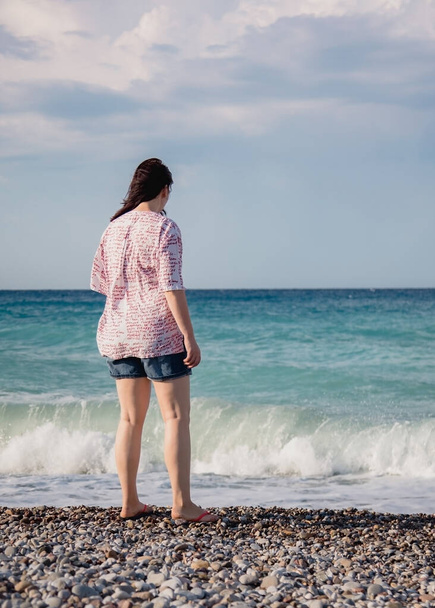 Młoda kobieta w spodenkach i koszuli stoi na żwirowej plaży i patrzy na fale Morza Śródziemnego, widok z tyłu, trzepotanie włosów na wietrze - Zdjęcie, obraz
