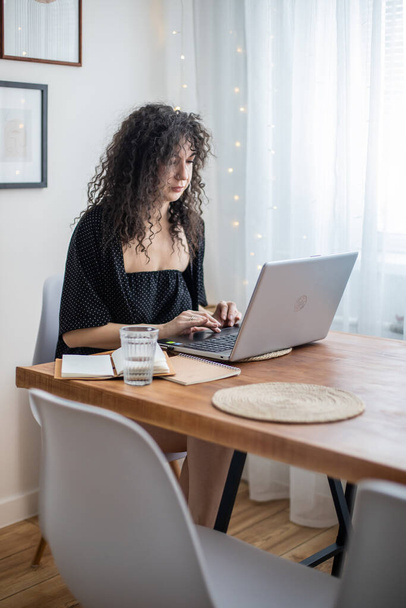 Lockige brünette freiberufliche Frau, die aus der Ferne arbeitet, benutzt Laptop im gemütlichen Heim skandinavischen Interieur - Foto, Bild