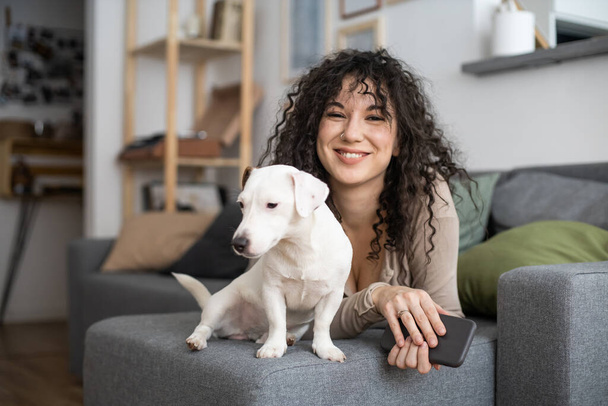 Cheerful curly woman kissing hugging little dog feeling love tenderness at cozy minimalistic home - Photo, Image