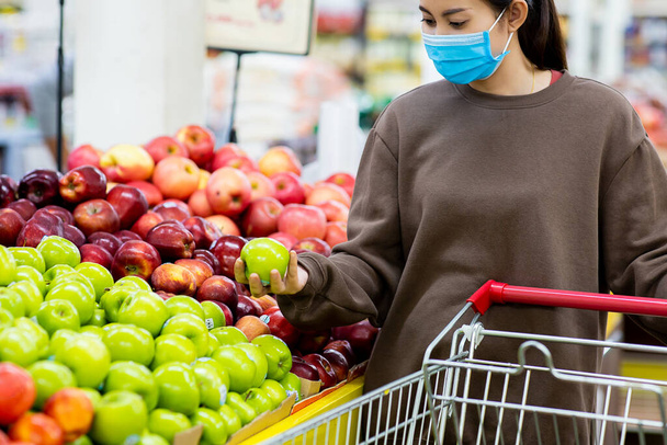 Jeune femme asiatique avec masque de protection poussant panier pour acheter des fruits frais au supermarché pendant l'épidémie de virus COVID-19. Concept de prévention COVID-19 Virus. - Photo, image