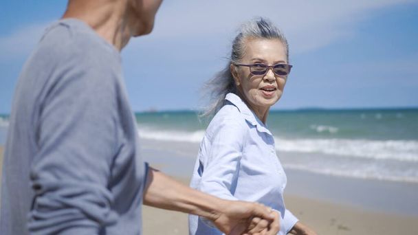 Happy Asian senior man and woman couple holding hands walking to the beach sunny with bright blue sky, Romantic elderly enjoy Travel summer vacation, plan life insurance at retirement couple concept - Photo, image