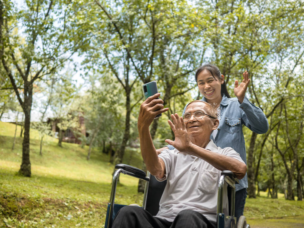 Grand-père en fauteuil roulant et petite-fille parler appel vidéo avec des parents via smartphone dans le parc. Vie de famille en vacances. - Photo, image