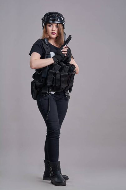 Uma jovem mulher está sentada com uma arma nas mãos em um fundo cinza - Foto, Imagem