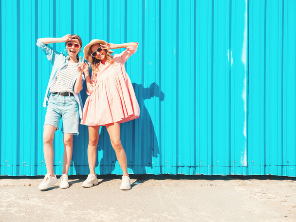 Two young beautiful smiling hipster female in trendy summer clothes and dress.Sexy carefree women posing in the street near blue wall.Positive pure models having fun at sunset, hugging and going crazy - Photo, image