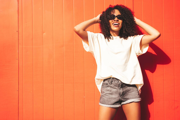 Beautiful black woman with afro curls hairstyle.Smiling hipster model in white t-shirt. Sexy carefree female posing in the street near red wall in sunglasses. Cheerful and happy - Valokuva, kuva