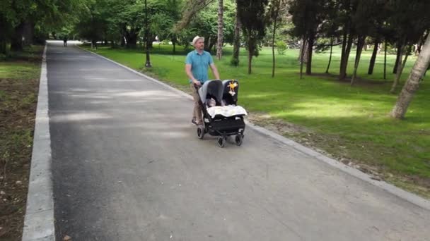 Padre con bambini in passeggino doppio in un parco. Uomo spingendo passeggino gemelli, passeggino. - Filmati, video