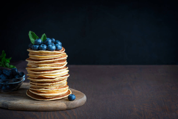 Stack of homemade american sweet pancakes or crepes decorated with fresh blueberries and mint leaves topping served on cutting board on dark brown wooden background. Image with copy space, horizontal - Фото, изображение